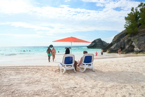A couple sits on beach chairs under a red umbrella on a Bermuda beach