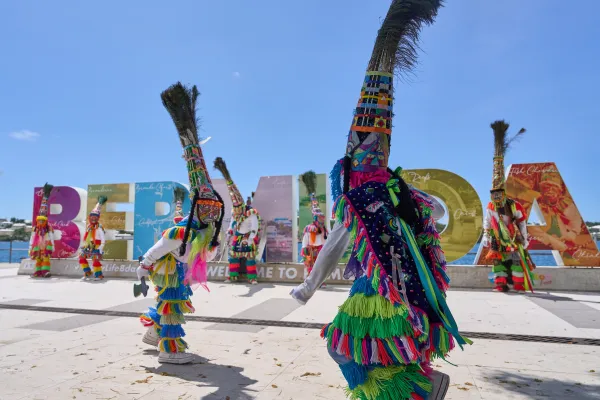 Gombey dancers in costume dancing in front of an outdoor Bermuda sign for Gombeys in the City