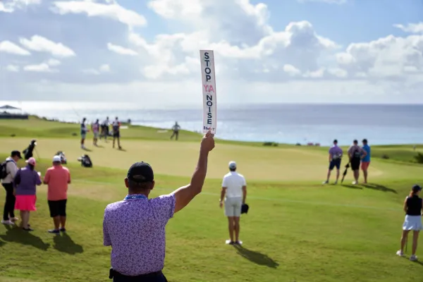 A golf course clerk holding a sign that reads "Stop Ya Noise" to quiet the crowd at the Butterfield Bermuda Championship PGA tour event