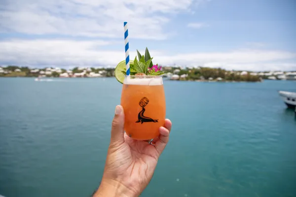 A person is holding a swizzle against a ocean backdrop.