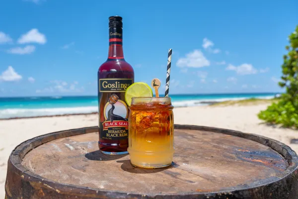 A view of a Goslings rum bottle with a dark n' stormy cocktail with a beach in the background.