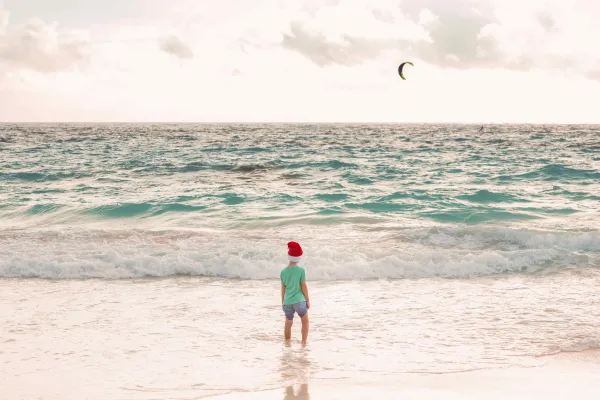 A little boy is standing on the beach in a Christmas hat.
