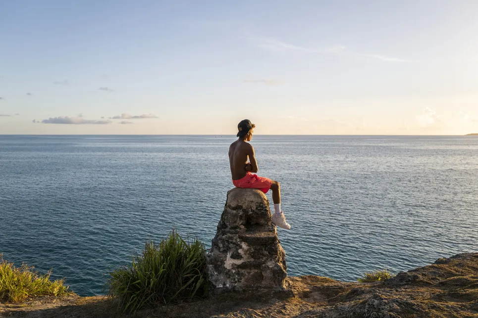 A young boy is sitting on a rock looking out at a calm ocean.