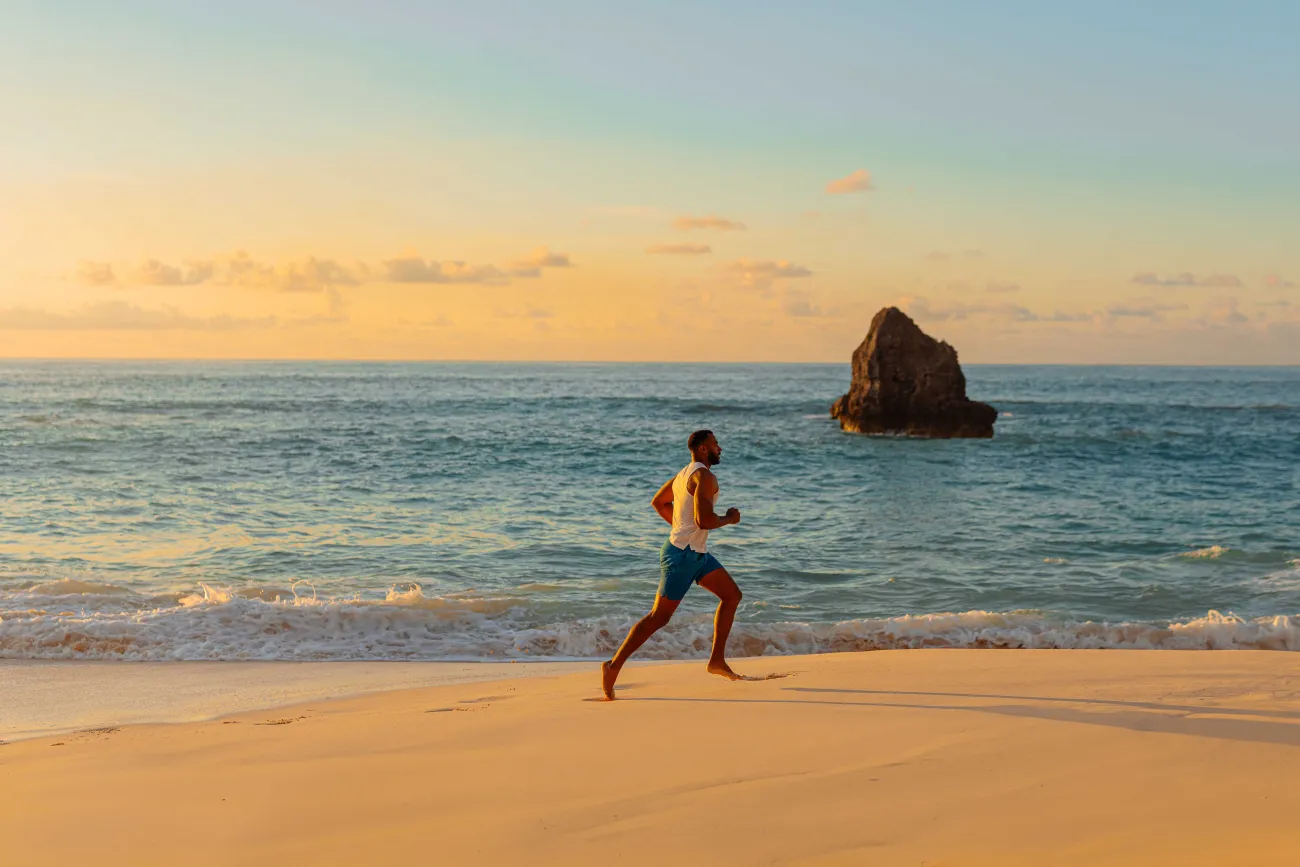A man is running at sunrise on a empty beach with calm ocean waters in the background.