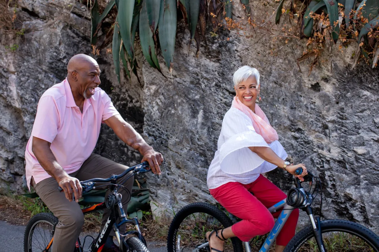 An older couple is riding bicycles and smiling at each other.