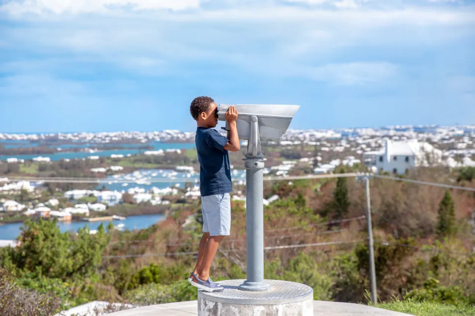 A little boy is looking through binoculars with a scenic background behind him.