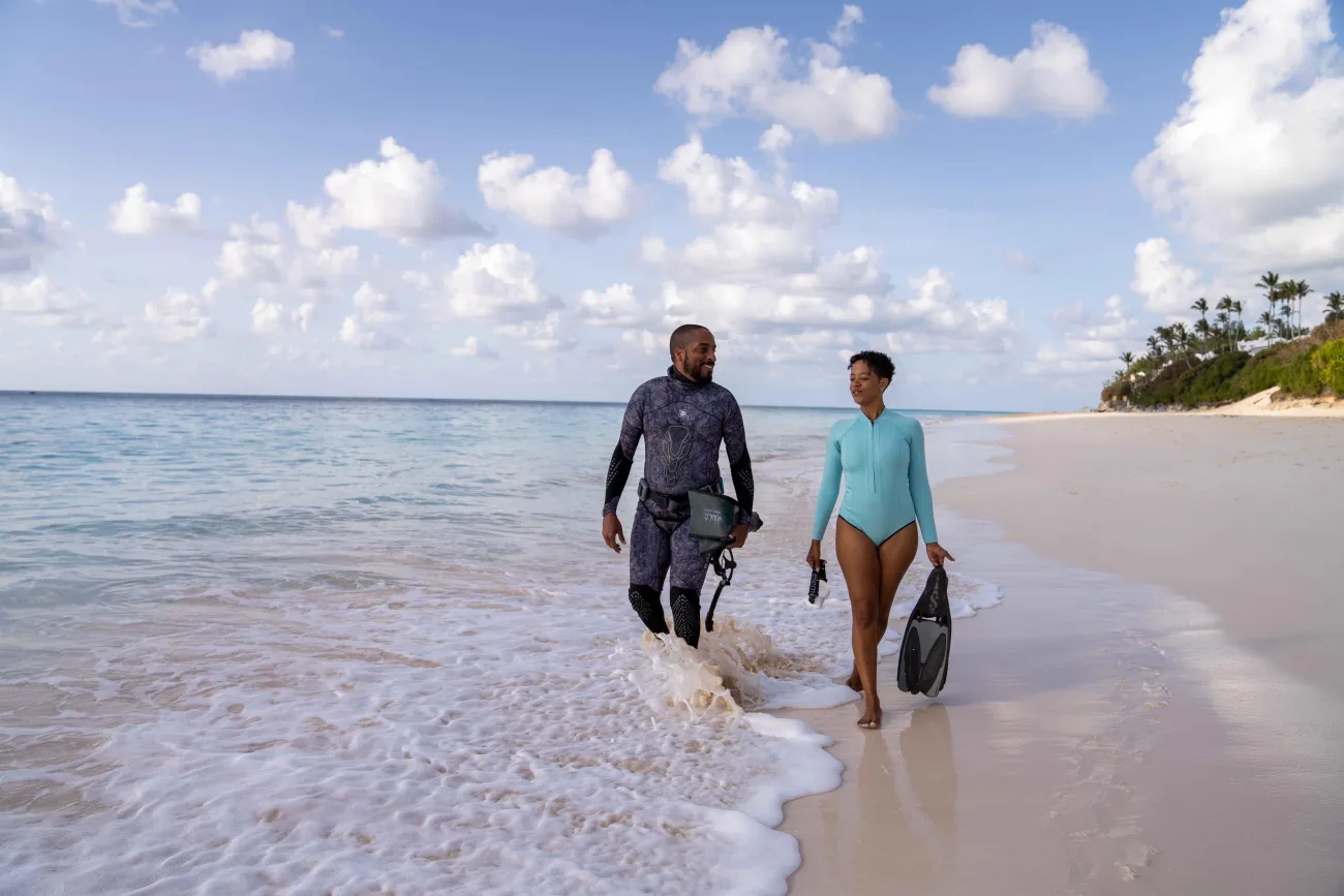 Two people are walking along the beach with scuba gear. 