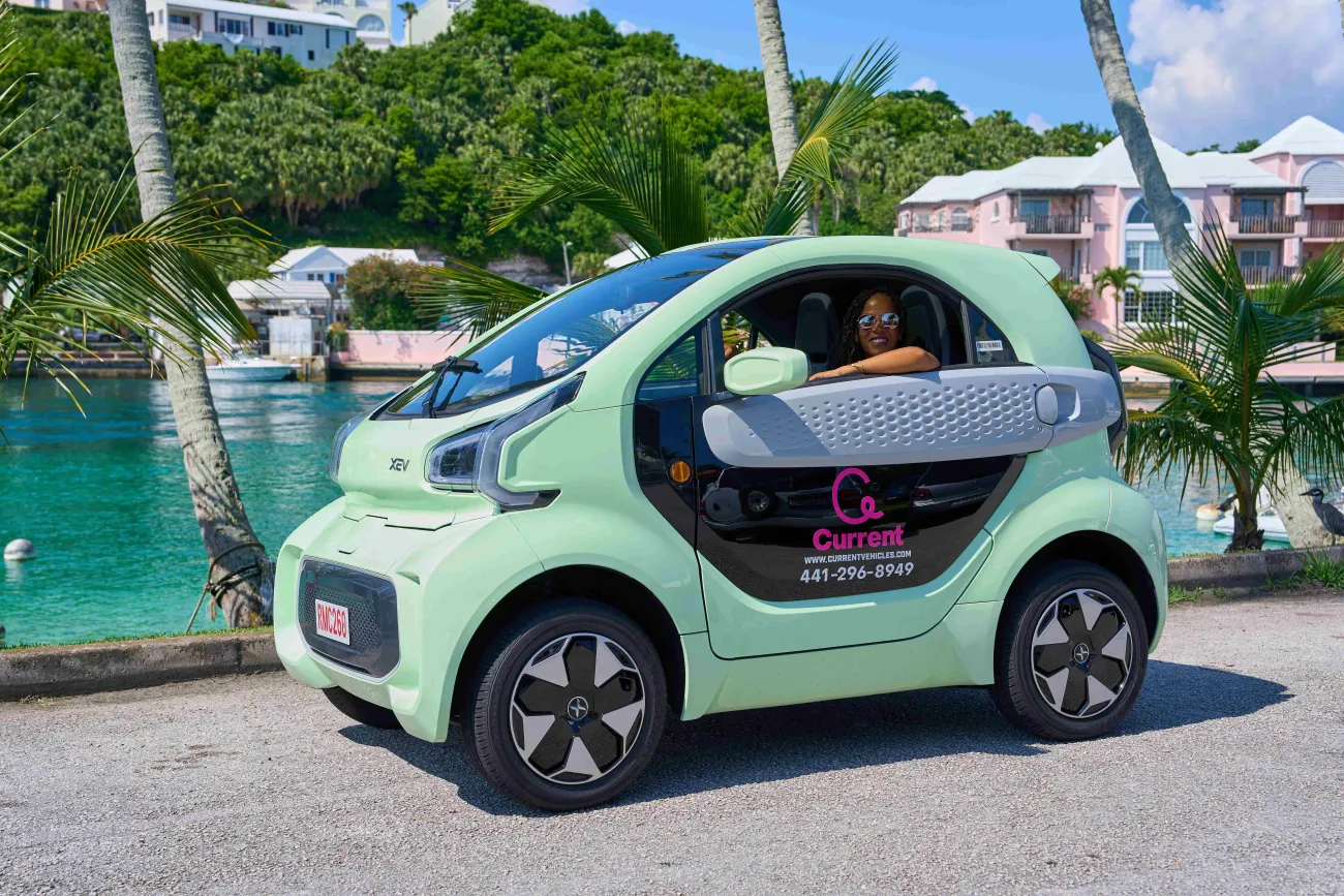 A woman is driving a microcar in scenic area with water and houses.