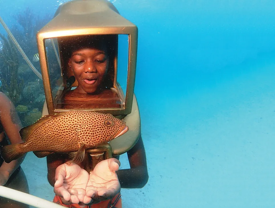 A young boy is wearing an underwater helmet looking at a fish.