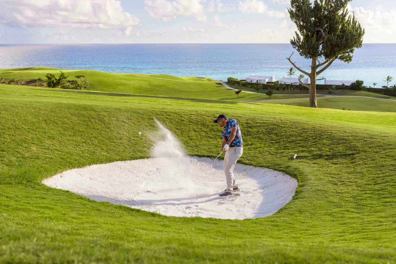 A man is hitting a golf ball out of a sand dune with the ocean in the background.