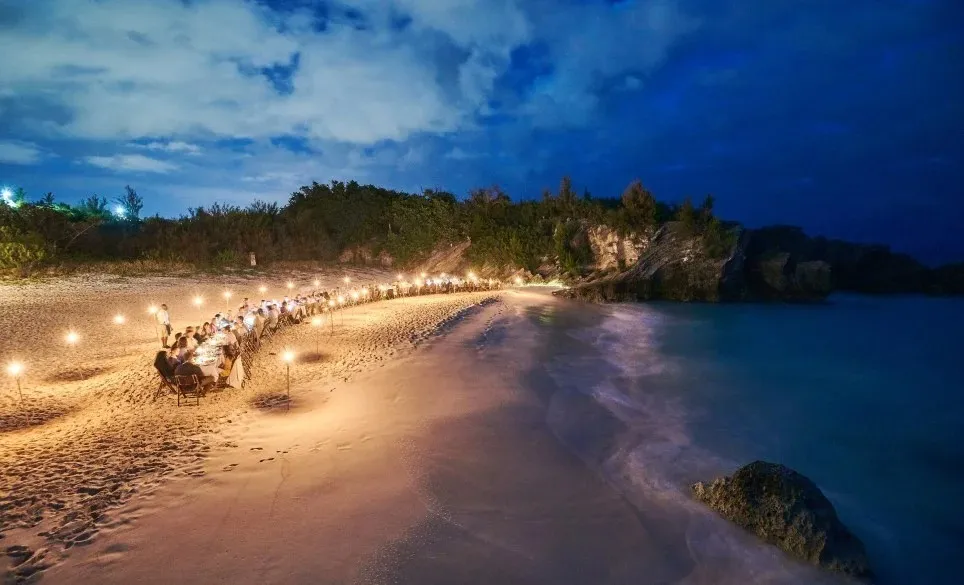Beach at night with a large table sitting people and lights surrounding them